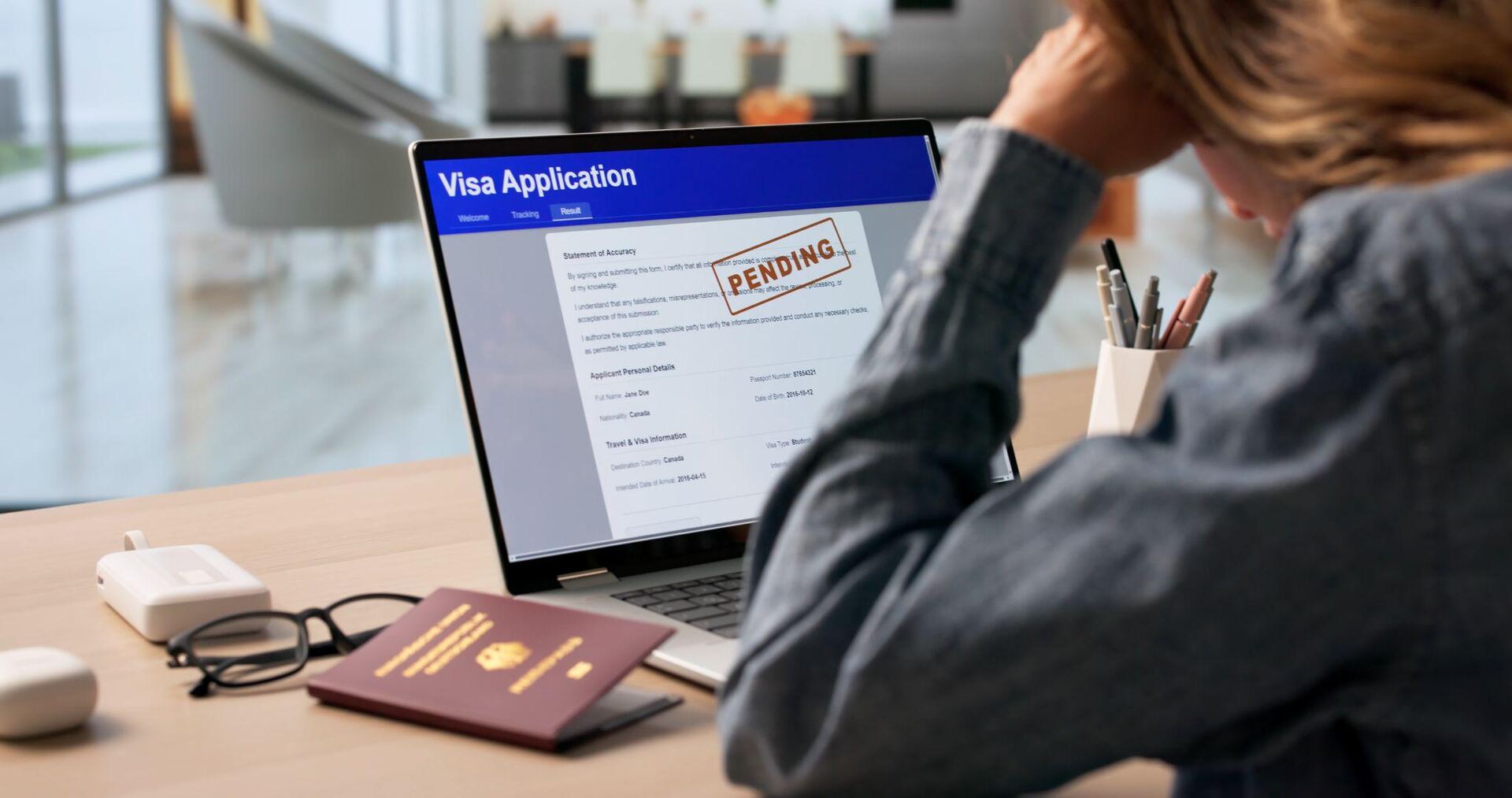 A worried applicant resting their head on one hand in front of a laptop showing a visa application stamped PENDING, with a passport and glasses on the desk.