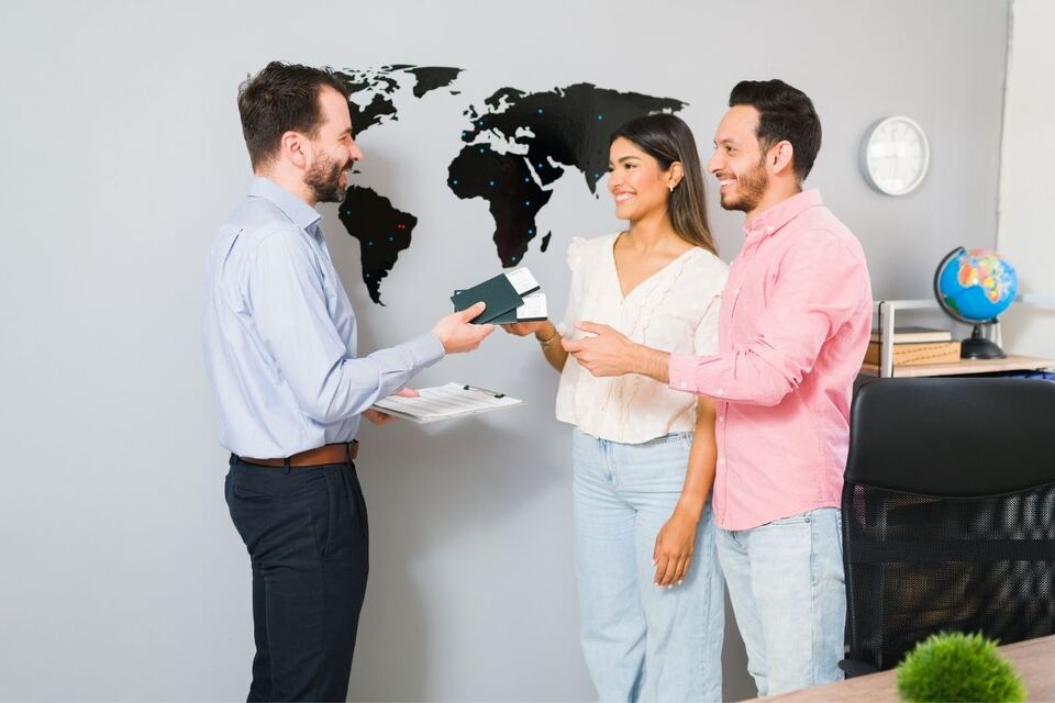 A visa consultant handing back two passports with documents tucked inside to a smiling couple, in an office with a world map on the wall.