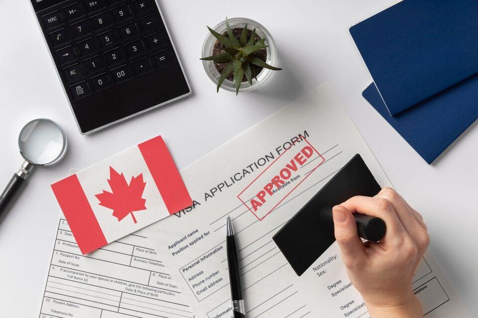 A Canada visa application form stamped "approved", with a Canadian flag, calculator, magnifying glass and passport folders on a white desk.