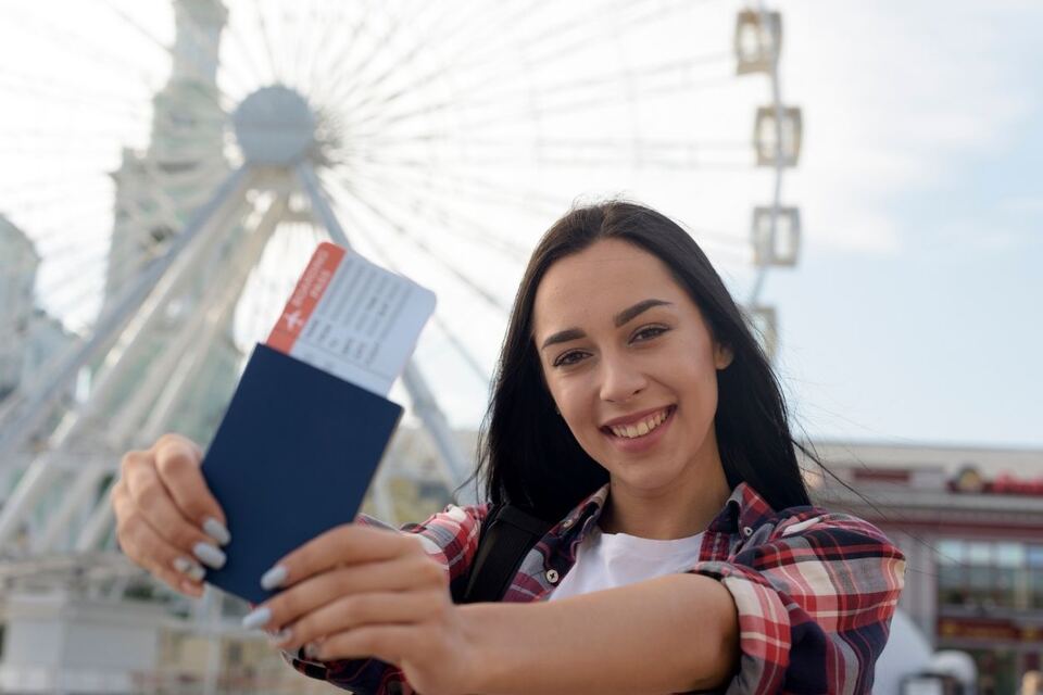 A smiling traveller holding up her passport with a boarding pass tucked inside, standing in front of a large ferris wheel.
