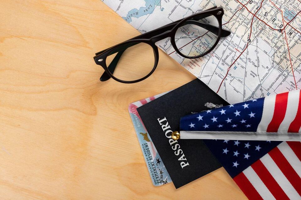 A passport, US permanent resident card and small American flag resting on a folded map beside a pair of glasses on a wooden desk.