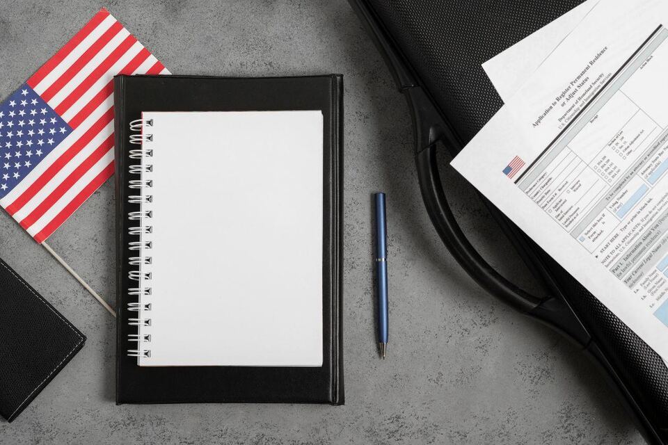 A flat-lay of a US visa application: a small American flag, a blank notebook and pen, a laptop bag and an immigration form on a grey desk.