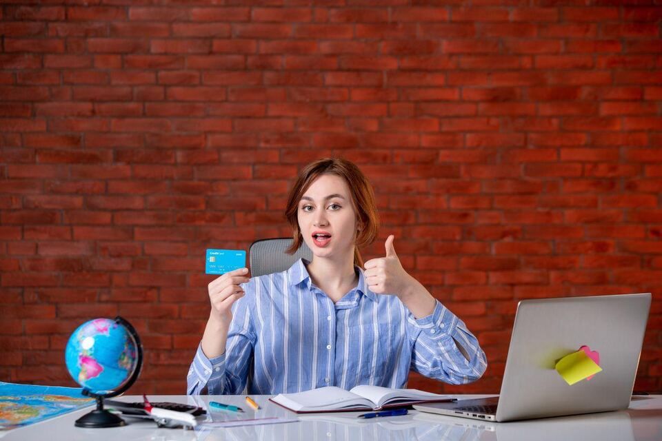 A woman at a desk holding a bank card and giving a thumbs up, with a globe, notebook and laptop beside her against a brick wall.