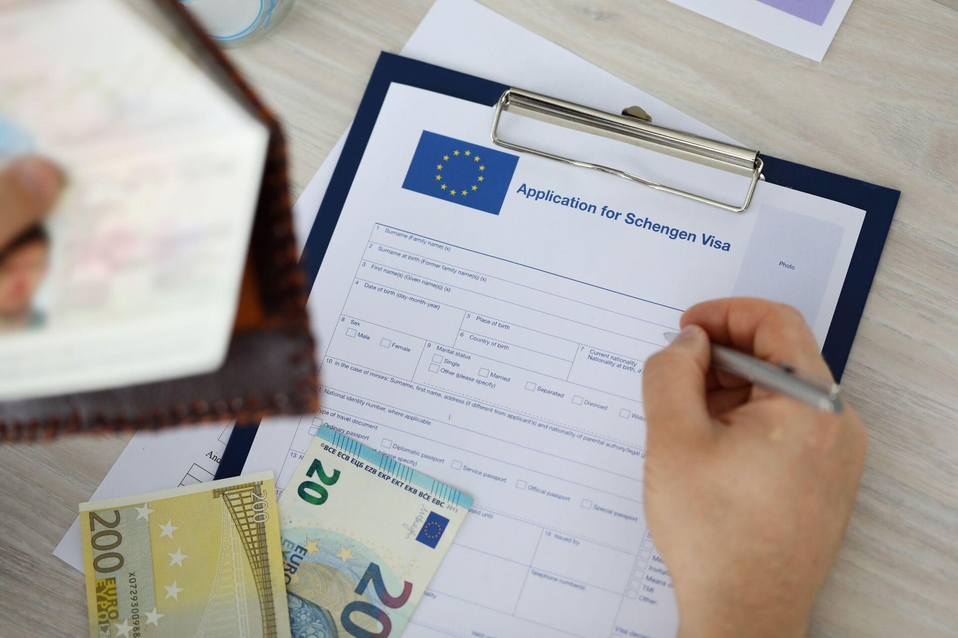 A hand filling in a Schengen visa application form on a clipboard, with 200 and 20 euro notes and an open passport beside it.