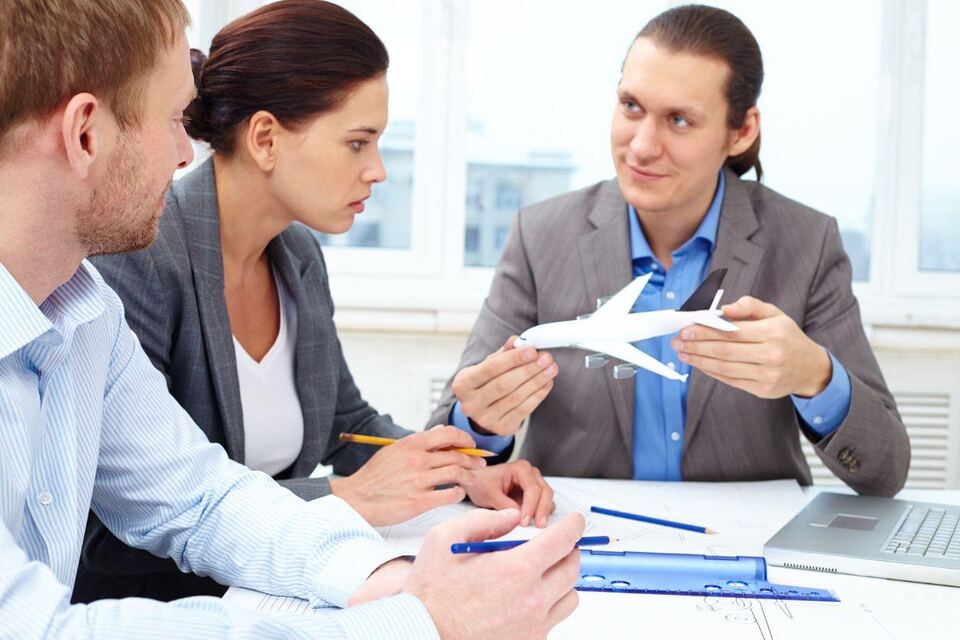Three consultants around a desk discussing travel plans, one holding a model aeroplane over an open notebook.