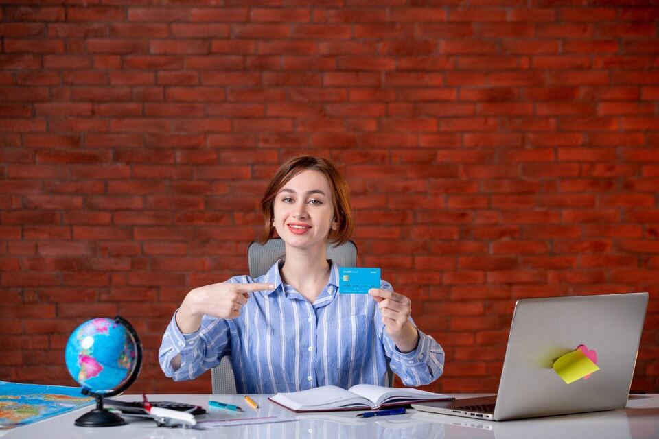A woman at a desk holding up a bank card beside a globe, notebook and laptop, planning the cost of a trip.