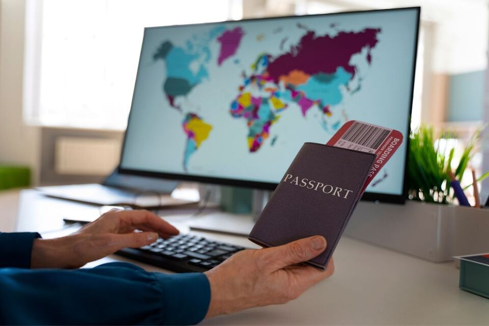 A traveller at a desk holding a passport with a boarding pass tucked inside, in front of a monitor showing a coloured world map.