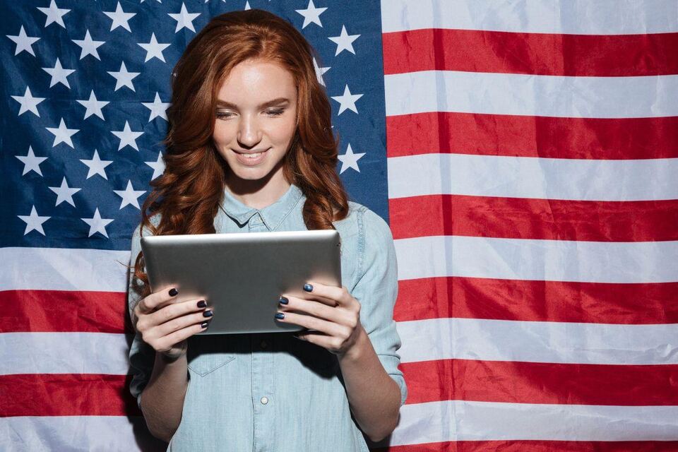 A woman smiling as she reads a tablet, standing in front of a large United States flag.